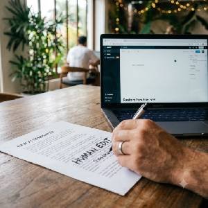 A stylized SEO featured image showing a close-up of a human hand on a laptop keyboard in a tropical café setting, humanizing a block of robotic text marked 'AI' by manual editing, with the Rankers Paradise brand theme.
